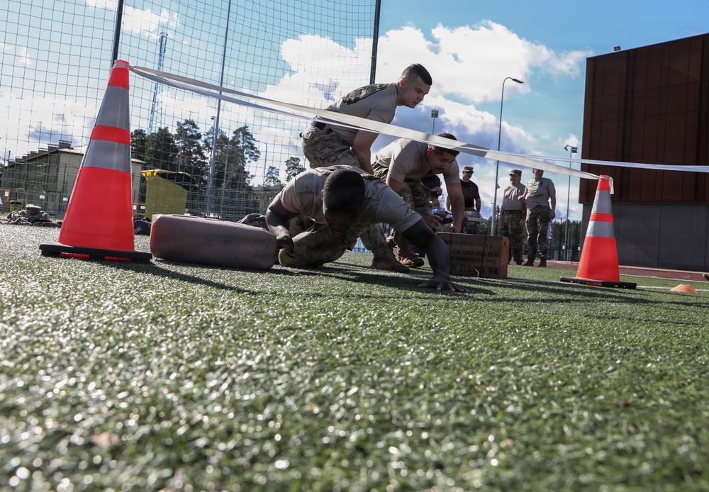 Military personnel are conducting a physical fitness or obstacle course exercise on an artificial turf field. Soldiers in camouflage uniforms are performing low crawl or push-up movements between orange traffic cones, while others observe in the background. The setting appears to be a military installation or training facility.