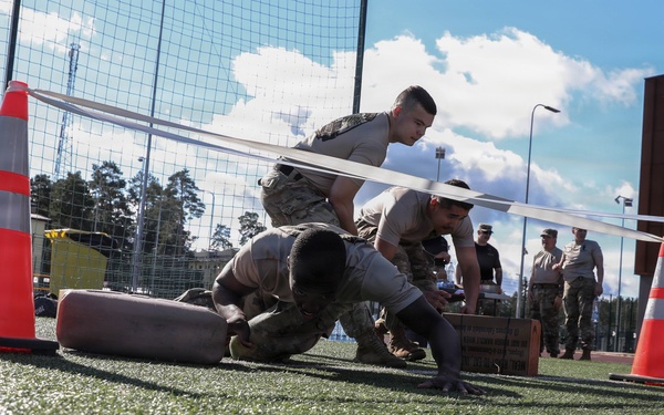 Military personnel are conducting a physical fitness or obstacle course exercise on an artificial turf field. Soldiers in camouflage uniforms are performing low crawl or push-up movements between orange traffic cones, while others observe in the background. The setting appears to be a military installation or training facility.