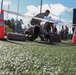 Military personnel are conducting a physical fitness or obstacle course exercise on an artificial turf field. Soldiers in camouflage uniforms are performing low crawl or push-up movements between orange traffic cones, while others observe in the background. The setting appears to be a military installation or training facility.