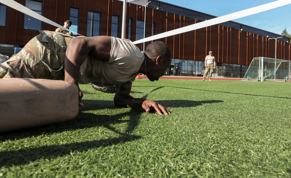 A soldier in military camouflage uniform and tan t-shirt performs a low crawl exercise on an artificial turf field, passing under a rope or tape barrier. The exercise appears to be part of a military fitness or obstacle course event conducted outdoors near a modern building. At least two other uniformed personnel are visible in the background, one appearing to observe or supervise.