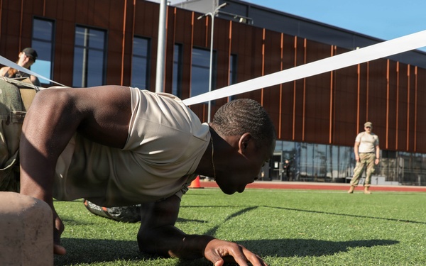 A soldier in military camouflage uniform and tan t-shirt performs a low crawl exercise on an artificial turf field, passing under a rope or tape barrier. The exercise appears to be part of a military fitness or obstacle course event conducted outdoors near a modern building. At least two other uniformed personnel are visible in the background, one appearing to observe or supervise.