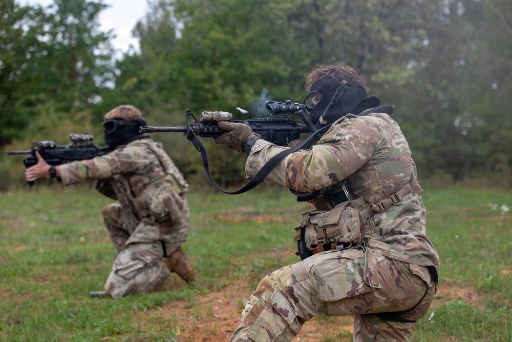 Two military personnel in full combat gear are in kneeling firing positions on an open grassy field. Both are wearing MultiCam/OCP camouflage uniforms and gas masks while actively firing rifles. Smoke is visible near the muzzle of the foreground weapon, indicating live or blank fire discharge.
