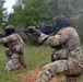 Two military personnel in full combat gear are in kneeling firing positions on an open grassy field. Both are wearing MultiCam/OCP camouflage uniforms and gas masks while actively firing rifles. Smoke is visible near the muzzle of the foreground weapon, indicating live or blank fire discharge.