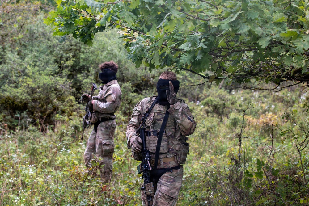 Two individuals wearing multicam camouflage uniforms and black balaclavas are positioned in a dense, green woodland or scrubland environment. Both are equipped with tactical gear including plate carriers, MOLLE pouches, and sling-mounted rifles. The scene appears consistent with a military exercise, airsoft/milsim event, or tactical training scenario.