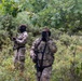 Two individuals wearing multicam camouflage uniforms and black balaclavas are positioned in a dense, green woodland or scrubland environment. Both are equipped with tactical gear including plate carriers, MOLLE pouches, and sling-mounted rifles. The scene appears consistent with a military exercise, airsoft/milsim event, or tactical training scenario.