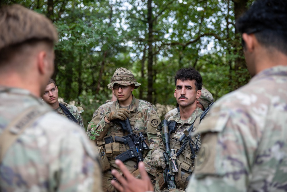 A group of soldiers in camouflage uniforms are gathered in a wooded outdoor setting, appearing to receive a briefing or instructions. Several personnel are armed with assault rifles and wearing tactical gear. The scene is consistent with a military training exercise or field operation.