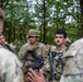 A group of soldiers in camouflage uniforms are gathered in a wooded outdoor setting, appearing to receive a briefing or instructions. Several personnel are armed with assault rifles and wearing tactical gear. The scene is consistent with a military training exercise or field operation.
