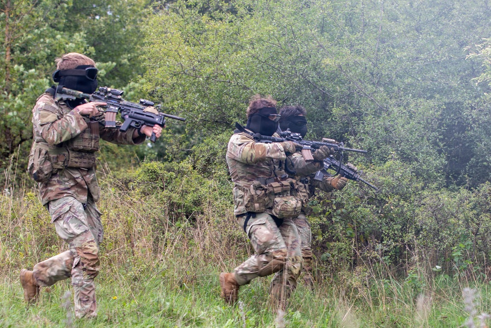 Two armed individuals wearing camouflage uniforms and black balaclavas are moving through a wooded, overgrown field while holding rifles in a ready position. Both are equipped with tactical gear including chest rigs and pouches. The setting appears to be a training exercise or military/airsoft simulation in a natural outdoor environment.