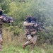 Two armed individuals wearing camouflage uniforms and black balaclavas are moving through a wooded, overgrown field while holding rifles in a ready position. Both are equipped with tactical gear including chest rigs and pouches. The setting appears to be a training exercise or military/airsoft simulation in a natural outdoor environment.