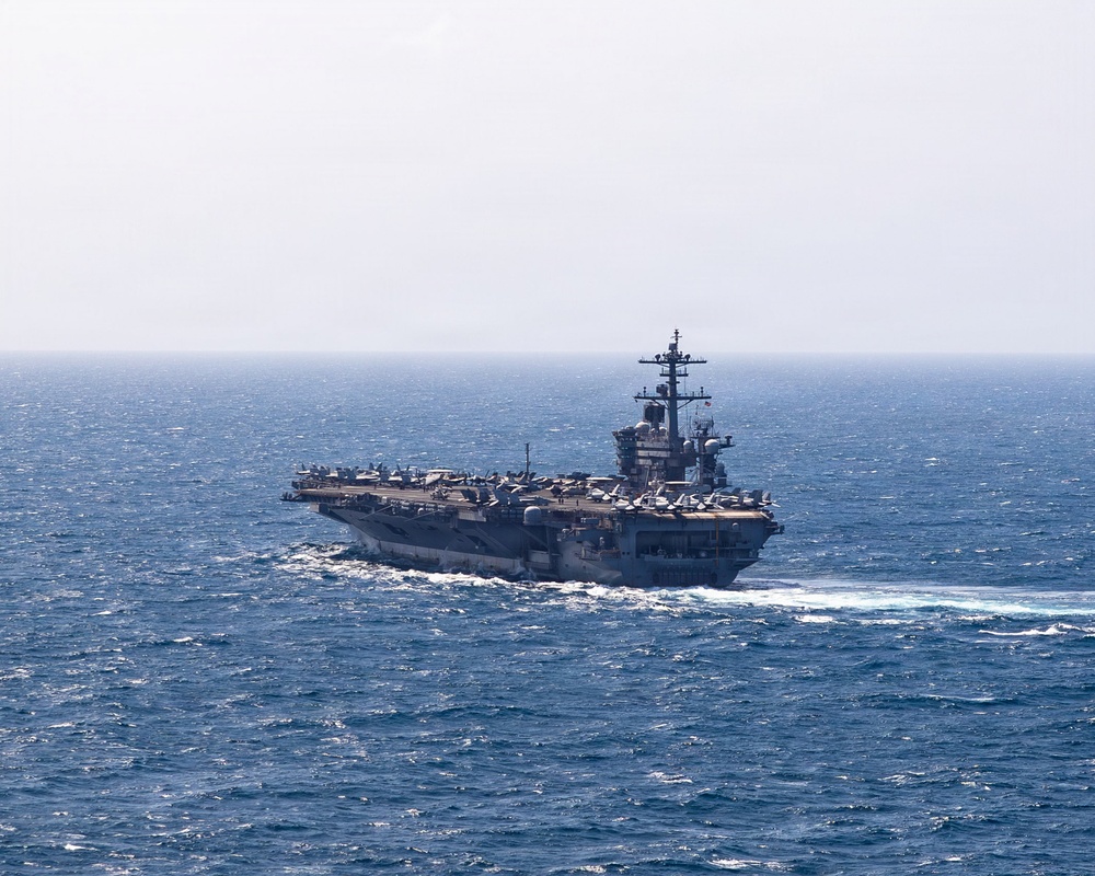 A large US Navy aircraft carrier is underway at sea, viewed from a slightly elevated angle. The flight deck is populated with multiple aircraft, and the ship is generating a visible wake as it moves through moderately choppy blue water. The island superstructure is visible on the starboard side with radar and communications equipment.