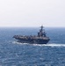 A large US Navy aircraft carrier is underway at sea, viewed from a slightly elevated angle. The flight deck is populated with multiple aircraft, and the ship is generating a visible wake as it moves through moderately choppy blue water. The island superstructure is visible on the starboard side with radar and communications equipment.
