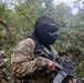 A person wearing MultiCam/OCP camouflage uniform with a US flag patch and a black face mask is holding an assault-style rifle in a wooded outdoor environment. A name tape reading 'BANDC__' is partially visible on the chest. Additional similarly equipped individuals are visible in the background, suggesting a group training exercise or airsoft/milsim event.