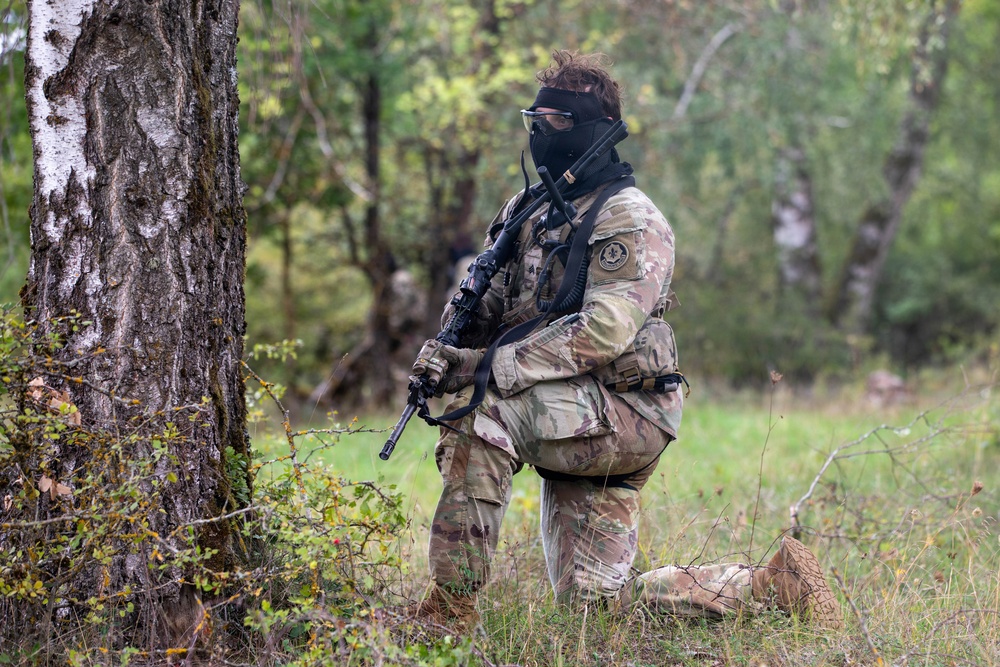A heavily equipped soldier or tactical operator in MultiCam/OCP camouflage uniform is kneeling beside a birch tree in a forested area. The individual is wearing a black balaclava and protective eyewear while holding an assault rifle. A second figure is faintly visible in the background, suggesting a small unit operation or training exercise.