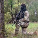 A heavily equipped soldier or tactical operator in MultiCam/OCP camouflage uniform is kneeling beside a birch tree in a forested area. The individual is wearing a black balaclava and protective eyewear while holding an assault rifle. A second figure is faintly visible in the background, suggesting a small unit operation or training exercise.