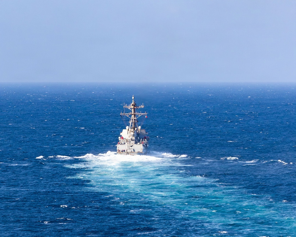 A naval warship, consistent with a U.S. Navy Arleigh Burke-class guided-missile destroyer, is photographed from astern while underway at sea. The vessel is moving at moderate to high speed, generating a prominent white wake and bow wash. The surrounding ocean is deep blue with choppy surface conditions under a hazy sky.