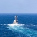 A naval warship, consistent with a U.S. Navy Arleigh Burke-class guided-missile destroyer, is photographed from astern while underway at sea. The vessel is moving at moderate to high speed, generating a prominent white wake and bow wash. The surrounding ocean is deep blue with choppy surface conditions under a hazy sky.