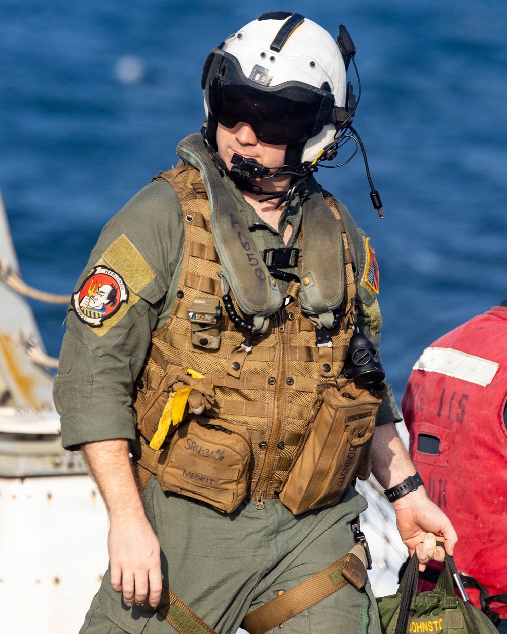 A military aviator or aircrew member is pictured in full flight gear near what appears to be a naval vessel with open water visible in the background. The individual is wearing a white flight helmet with visor, olive drab flight suit, and a tan tactical survival vest with multiple pouches. Visible markings and patches suggest US Navy affiliation.