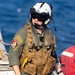 A military aviator or aircrew member is pictured in full flight gear near what appears to be a naval vessel with open water visible in the background. The individual is wearing a white flight helmet with visor, olive drab flight suit, and a tan tactical survival vest with multiple pouches. Visible markings and patches suggest US Navy affiliation.
