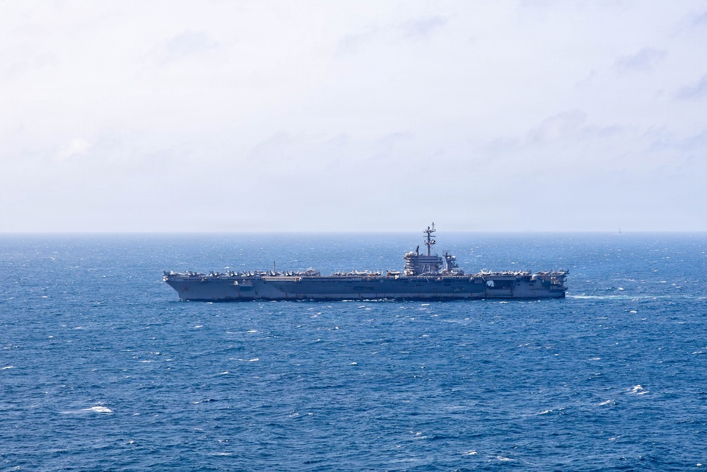 A large aircraft carrier is underway at sea, viewed from a distance in a broadside profile. The vessel appears to be a Nimitz-class or similar supercarrier, with a flat flight deck visible and an island superstructure on the starboard side. The ship is sailing through moderately choppy blue ocean waters under an overcast sky.