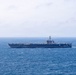 A large aircraft carrier is underway at sea, viewed from a distance in a broadside profile. The vessel appears to be a Nimitz-class or similar supercarrier, with a flat flight deck visible and an island superstructure on the starboard side. The ship is sailing through moderately choppy blue ocean waters under an overcast sky.
