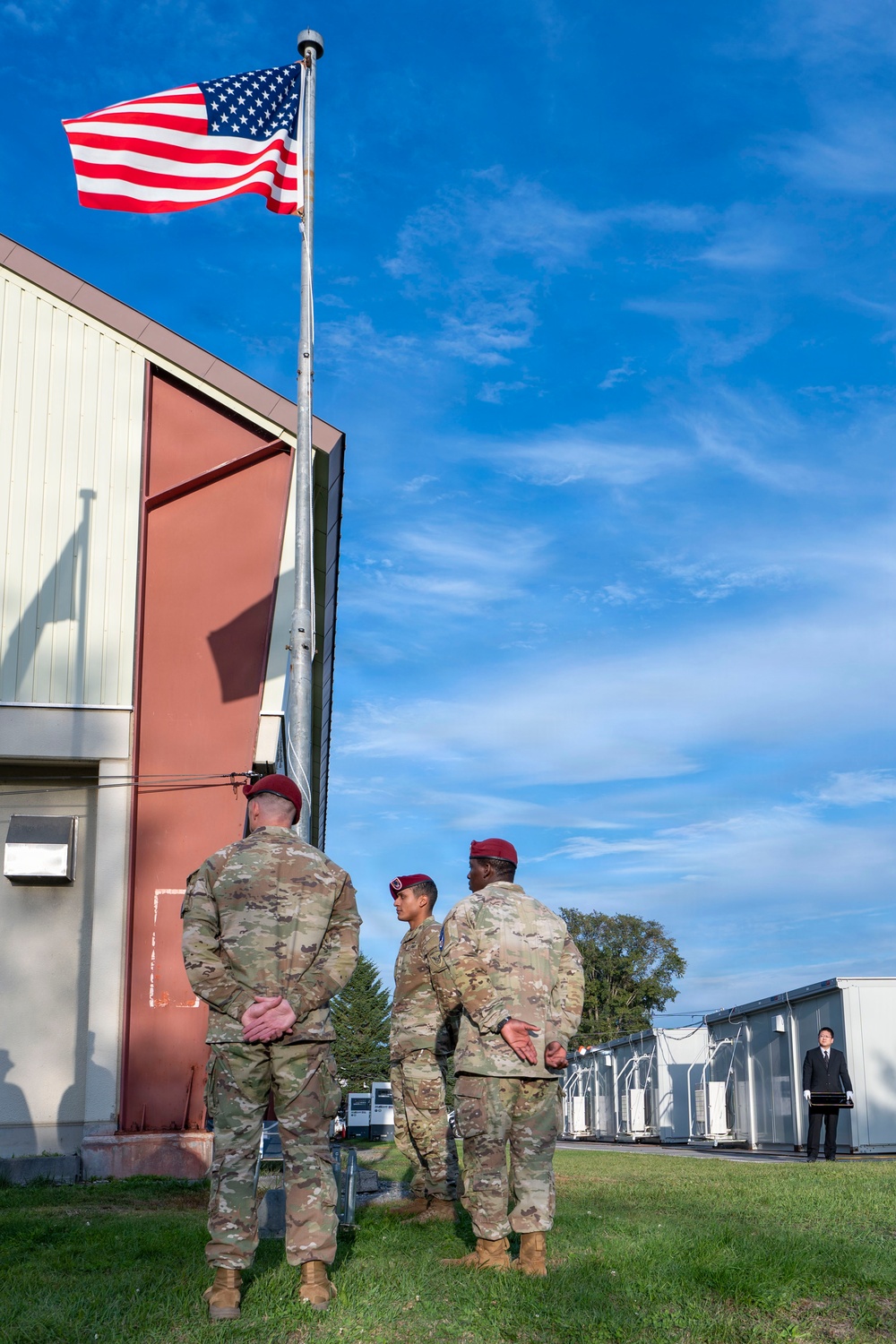 Three U.S. Army soldiers wearing MultiCam (OCP) uniforms and maroon berets stand near a flagpole flying the American flag at what appears to be a military installation. A civilian in dark attire stands in the background to the right. The scene suggests an informal gathering or conversation near a flag formation area.