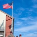 Three U.S. Army soldiers wearing MultiCam (OCP) uniforms and maroon berets stand near a flagpole flying the American flag at what appears to be a military installation. A civilian in dark attire stands in the background to the right. The scene suggests an informal gathering or conversation near a flag formation area.