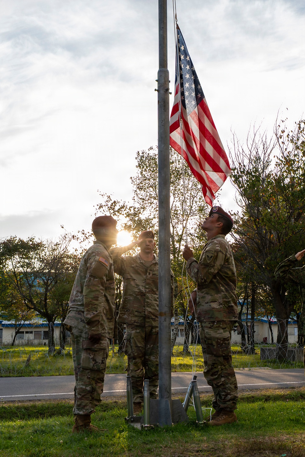 Three U.S. Army soldiers in OCP (Operational Camouflage Pattern) uniforms are raising an American flag on a portable flagpole during what appears to be an early morning or evening ceremony. The soldiers are wearing maroon/red berets, indicating possible airborne or special operations affiliation. The scene is set on a military installation with trees and low buildings visible in the background.