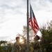Three U.S. Army soldiers in OCP (Operational Camouflage Pattern) uniforms are raising an American flag on a portable flagpole during what appears to be an early morning or evening ceremony. The soldiers are wearing maroon/red berets, indicating possible airborne or special operations affiliation. The scene is set on a military installation with trees and low buildings visible in the background.