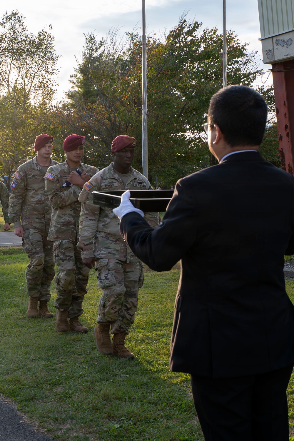 A military ceremony is taking place outdoors, likely an awards or decoration ceremony. Three U.S. Army soldiers wearing maroon berets and MultiCam (OCP) uniforms are standing in formation while a civilian in a dark suit, wearing white gloves, holds a presentation tray toward them. The setting appears to be a military installation with trees showing early autumn foliage in the background.