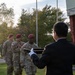 A military ceremony is taking place outdoors, likely an awards or decoration ceremony. Three U.S. Army soldiers wearing maroon berets and MultiCam (OCP) uniforms are standing in formation while a civilian in a dark suit, wearing white gloves, holds a presentation tray toward them. The setting appears to be a military installation with trees showing early autumn foliage in the background.