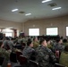 A large military briefing or conference is taking place in a formal meeting room, with personnel from multiple nations wearing various camouflage uniforms seated in rows of chairs. A presenter stands at a podium at the front of the room, with three projection screens displaying content behind them. Attendees appear to be actively engaged, with some using laptops.