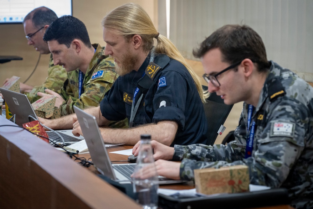 Four military personnel are seated at a conference table working on laptops during what appears to be a joint military exercise or briefing. The group includes members from at least two nations — Australia and New Zealand — identifiable by flag patches and uniform styles. They appear engaged in active work or coordination tasks.