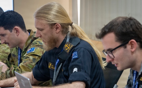 Four military personnel are seated at a conference table working on laptops during what appears to be a joint military exercise or briefing. The group includes members from at least two nations — Australia and New Zealand — identifiable by flag patches and uniform styles. They appear engaged in active work or coordination tasks.