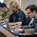 Four military personnel are seated at a conference table working on laptops during what appears to be a joint military exercise or briefing. The group includes members from at least two nations — Australia and New Zealand — identifiable by flag patches and uniform styles. They appear engaged in active work or coordination tasks.