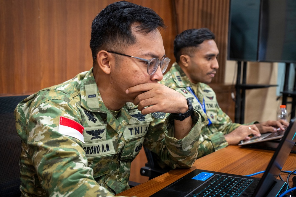 Two Indonesian military personnel in camouflage uniforms are seated at a wooden desk working on laptops. The foreground soldier, identified by name tape 'GROHO A.W' and 'TNI A' branch insignia, is focused intently on his screen with his hand raised to his chin in a contemplative pose. Both soldiers wear Indonesian flag patches and appear to be engaged in a training or operational briefing environment.