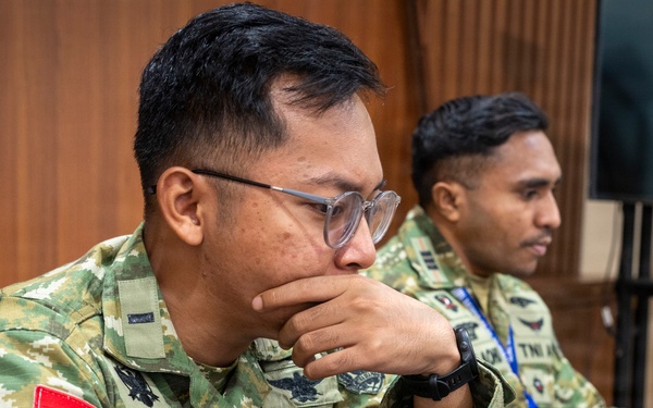 Two Indonesian military personnel in camouflage uniforms are seated at a wooden desk working on laptops. The foreground soldier, identified by name tape 'GROHO A.W' and 'TNI A' branch insignia, is focused intently on his screen with his hand raised to his chin in a contemplative pose. Both soldiers wear Indonesian flag patches and appear to be engaged in a training or operational briefing environment.