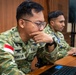 Two Indonesian military personnel in camouflage uniforms are seated at a wooden desk working on laptops. The foreground soldier, identified by name tape 'GROHO A.W' and 'TNI A' branch insignia, is focused intently on his screen with his hand raised to his chin in a contemplative pose. Both soldiers wear Indonesian flag patches and appear to be engaged in a training or operational briefing environment.