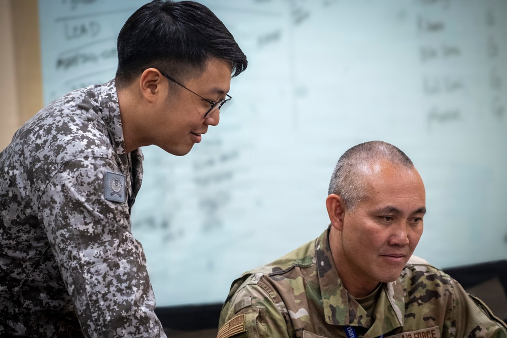 Two military personnel in different camouflage uniforms are collaborating over what appears to be a document or screen. One individual is leaning over while the other is seated, both appearing engaged in a briefing or planning session. A whiteboard with handwritten notes is visible in the background.