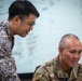 Two military personnel in different camouflage uniforms are collaborating over what appears to be a document or screen. One individual is leaning over while the other is seated, both appearing engaged in a briefing or planning session. A whiteboard with handwritten notes is visible in the background.