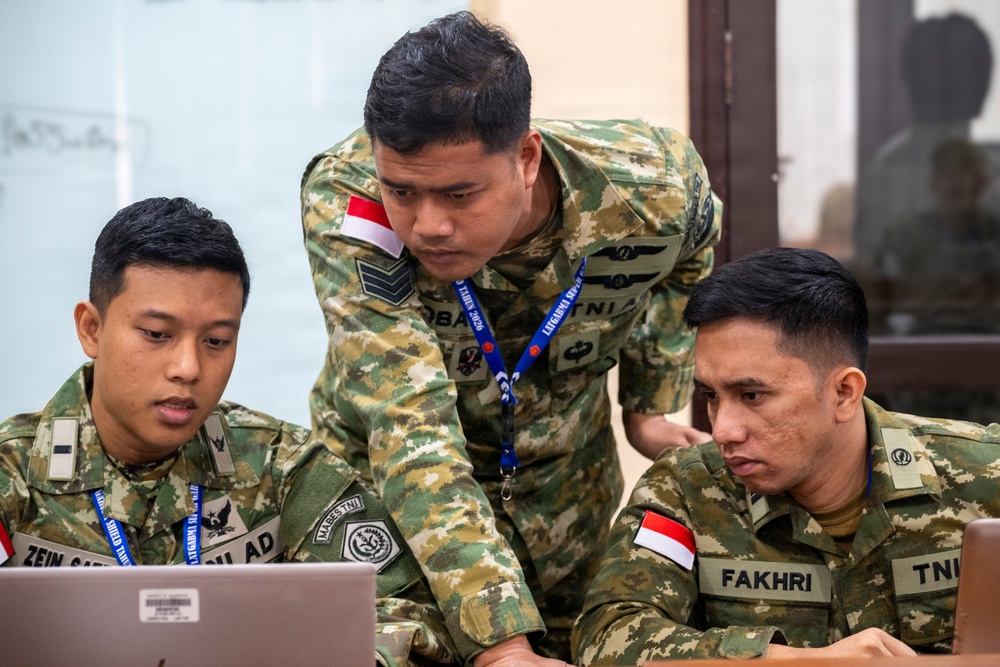 Three Indonesian military personnel in camouflage uniforms are gathered around a laptop computer in what appears to be an indoor briefing or operations room. The soldiers are focused on the screen, with one standing and pointing while the other two are seated. Name tags and Indonesian flag patches are visible on their uniforms.