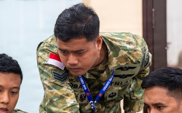 Three Indonesian military personnel in camouflage uniforms are gathered around a laptop computer in what appears to be an indoor briefing or operations room. The soldiers are focused on the screen, with one standing and pointing while the other two are seated. Name tags and Indonesian flag patches are visible on their uniforms.