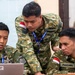 Three Indonesian military personnel in camouflage uniforms are gathered around a laptop computer in what appears to be an indoor briefing or operations room. The soldiers are focused on the screen, with one standing and pointing while the other two are seated. Name tags and Indonesian flag patches are visible on their uniforms.