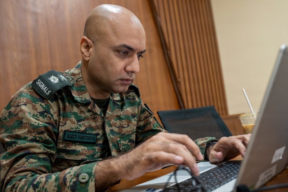A military personnel member in camouflage uniform is focused on working at a laptop computer. The individual's name tape reads 'AMIT VASUDEVA' and the shoulder patch indicates 'SIGNALS' branch. The setting appears to be an indoor workspace with wooden paneling in the background.