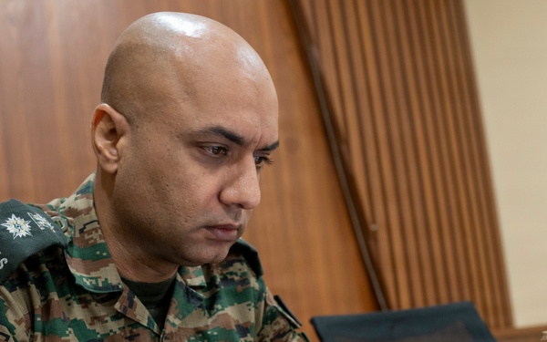 A military personnel member in camouflage uniform is focused on working at a laptop computer. The individual's name tape reads 'AMIT VASUDEVA' and the shoulder patch indicates 'SIGNALS' branch. The setting appears to be an indoor workspace with wooden paneling in the background.