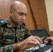 A military personnel member in camouflage uniform is focused on working at a laptop computer. The individual's name tape reads 'AMIT VASUDEVA' and the shoulder patch indicates 'SIGNALS' branch. The setting appears to be an indoor workspace with wooden paneling in the background.