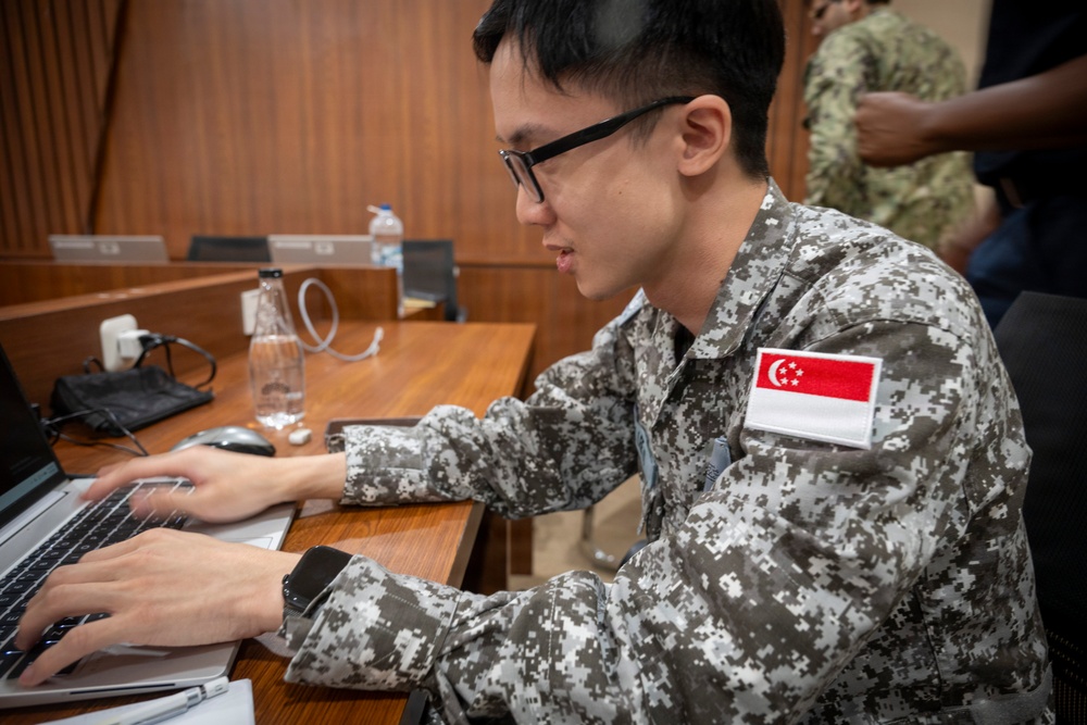 A military personnel wearing a Singapore Armed Forces uniform is working on a laptop computer at a wooden desk. The soldier is identifiable by the Singapore flag patch on the right sleeve of their digital camouflage uniform. The setting appears to be an indoor briefing room or conference facility.