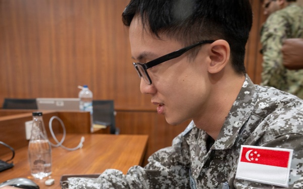 A military personnel wearing a Singapore Armed Forces uniform is working on a laptop computer at a wooden desk. The soldier is identifiable by the Singapore flag patch on the right sleeve of their digital camouflage uniform. The setting appears to be an indoor briefing room or conference facility.
