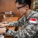 A military personnel wearing a Singapore Armed Forces uniform is working on a laptop computer at a wooden desk. The soldier is identifiable by the Singapore flag patch on the right sleeve of their digital camouflage uniform. The setting appears to be an indoor briefing room or conference facility.