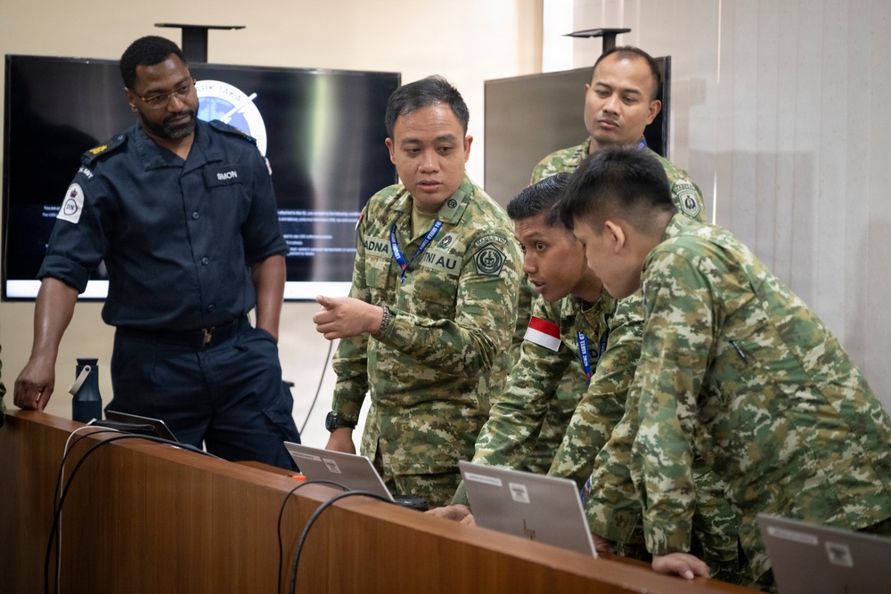 A group of military personnel from multiple nations are gathered around laptops on a wooden table in what appears to be a briefing or operations room. One soldier in camouflage uniform is pointing at a laptop screen while others lean in to observe, suggesting a collaborative training or planning exercise. A large display screen showing a logo or emblem is visible in the background.