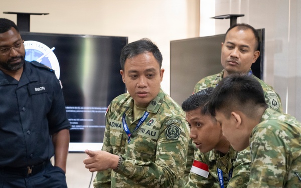 A group of military personnel from multiple nations are gathered around laptops on a wooden table in what appears to be a briefing or operations room. One soldier in camouflage uniform is pointing at a laptop screen while others lean in to observe, suggesting a collaborative training or planning exercise. A large display screen showing a logo or emblem is visible in the background.